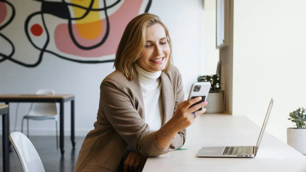 Uśmiechnięta kobieta w beżowym żakiecie korzysta z telefonu komórkowego i laptopa w jasnym biurze z abstrakcyjnym obrazem w tle, porównując ceny usług internetowych w 2024 roku.
