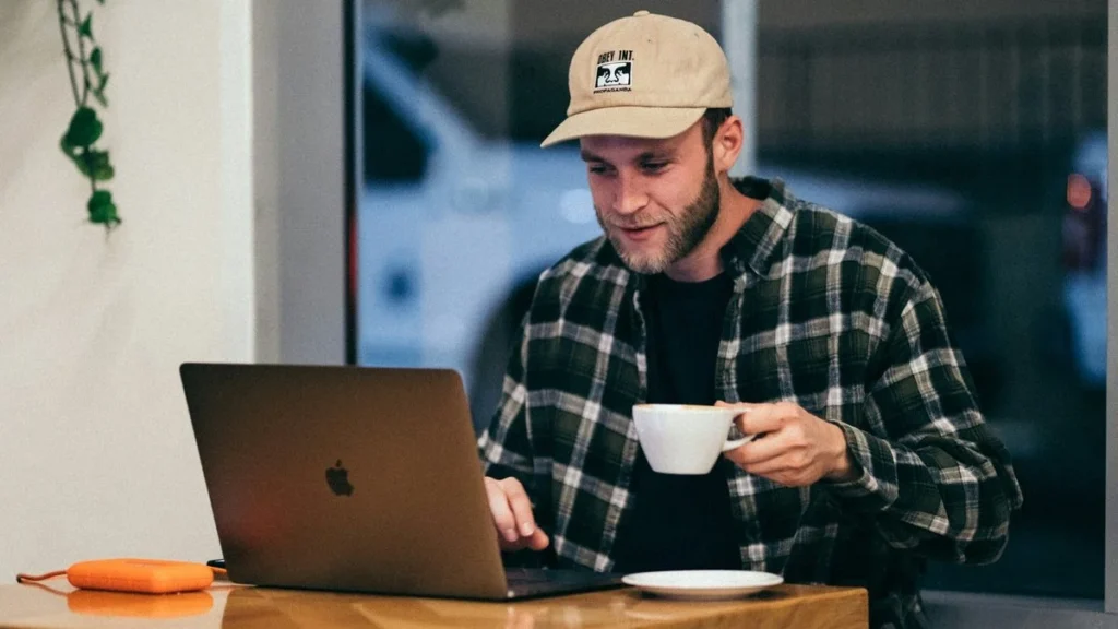 Młody mężczyzna w kratowanej koszuli i beżowej czapce siedzi w kawiarni i korzysta z laptopa Apple, popijając kawę, podczas przeglądania informacji o cenach projektowania stron internetowych w 2024 roku.